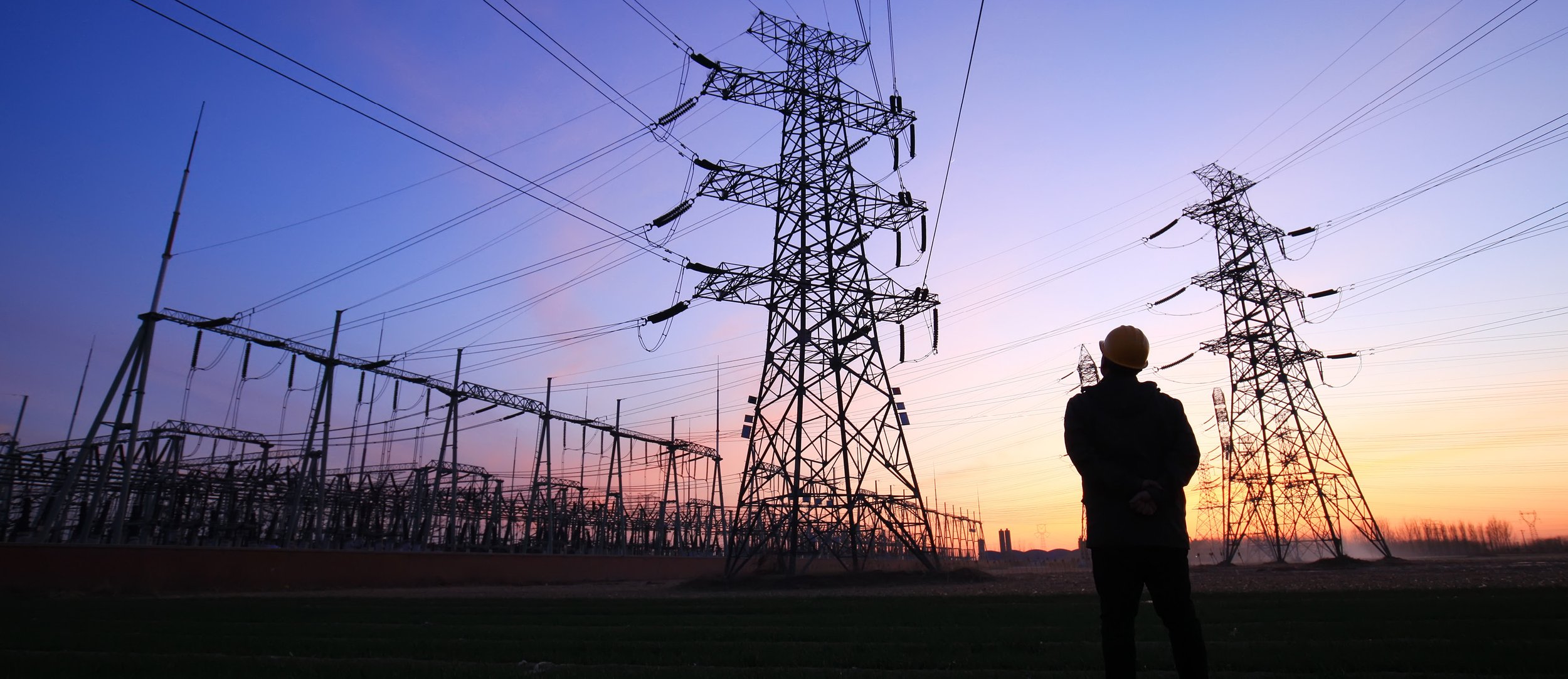 A person wearing a safety helmet standing and observing electrical power lines and towers during sunset.
