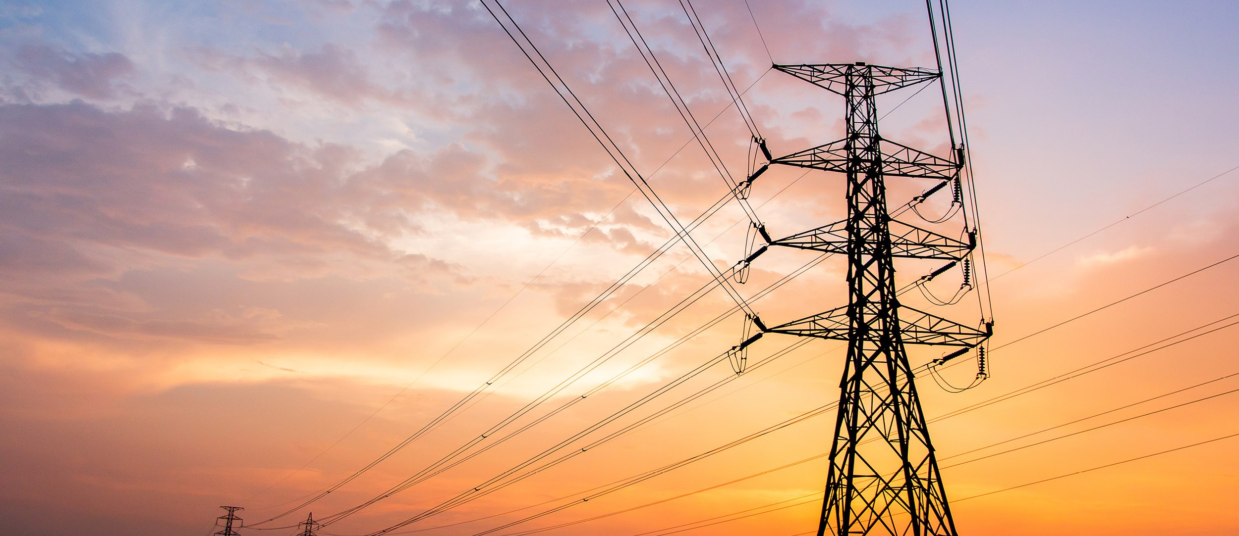 A power line tower with multiple electrical wires against a colorful sunset sky with clouds.