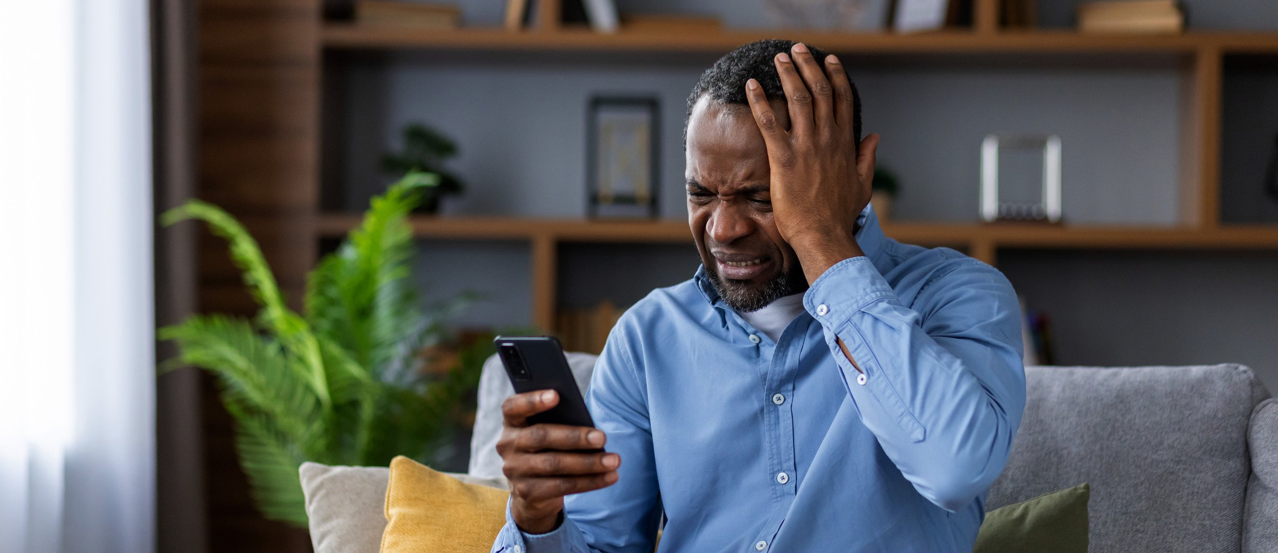 Man with short curly hair and a beard sitting on a sofa, holding a smartphone in his right hand and touching his head with his left hand, looking distressed.