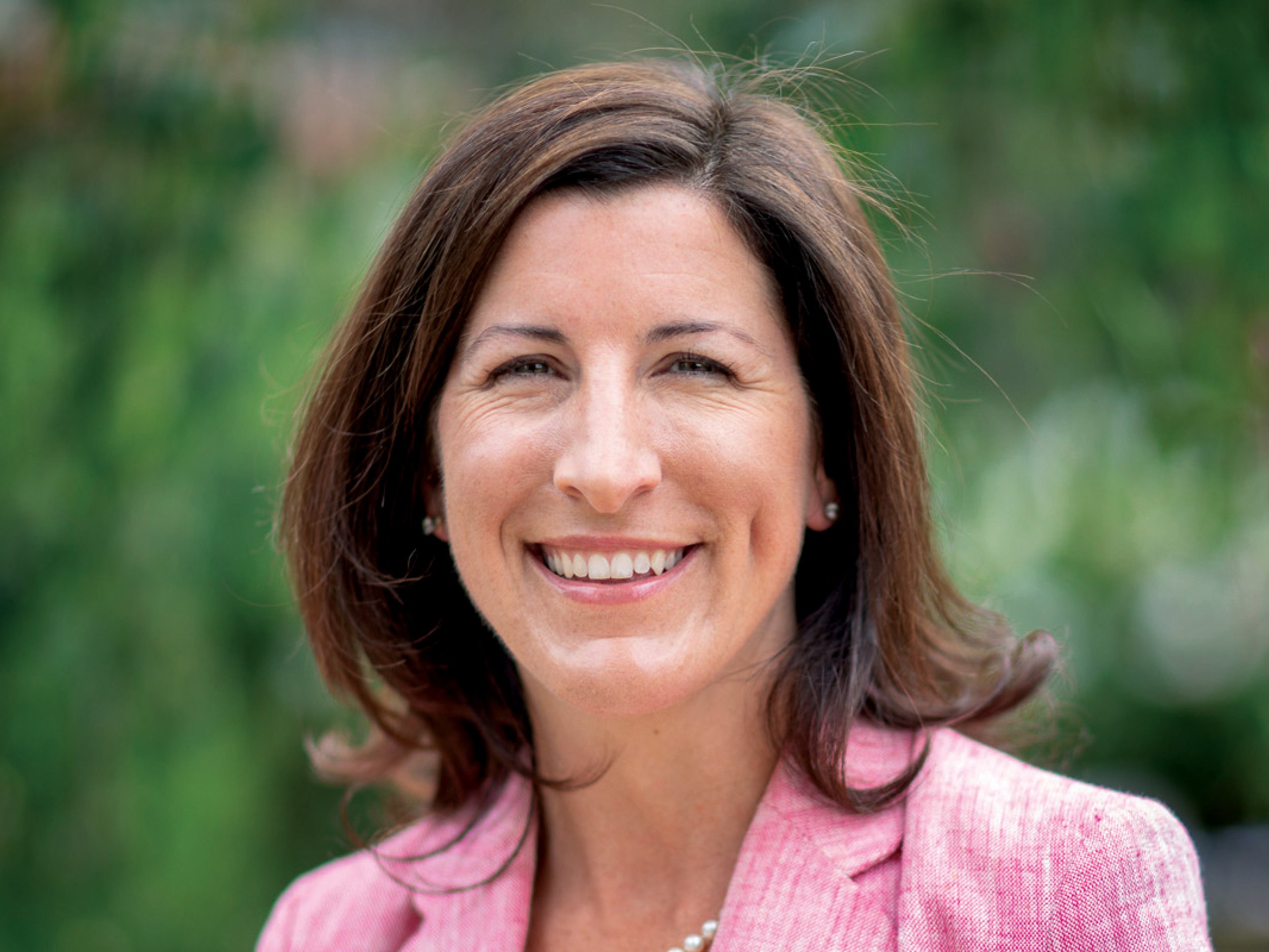A woman with shoulder-length brown hair, smiling, wearing a pink blazer and pearl earrings outdoors with a blurred green background.