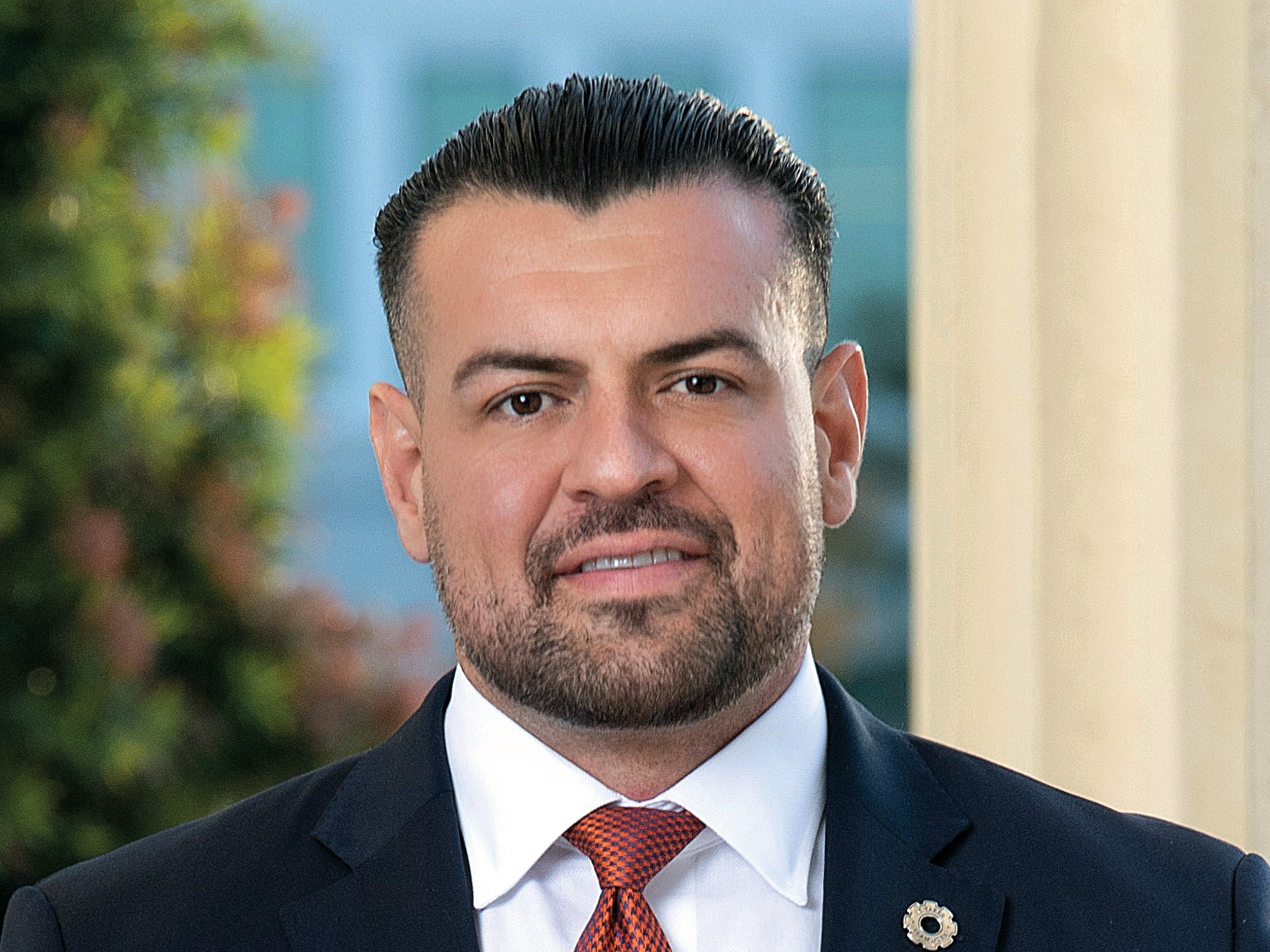 Headshot of a man in a suit with a white shirt and orange tie, outdoors with blurred greenery and building background.
