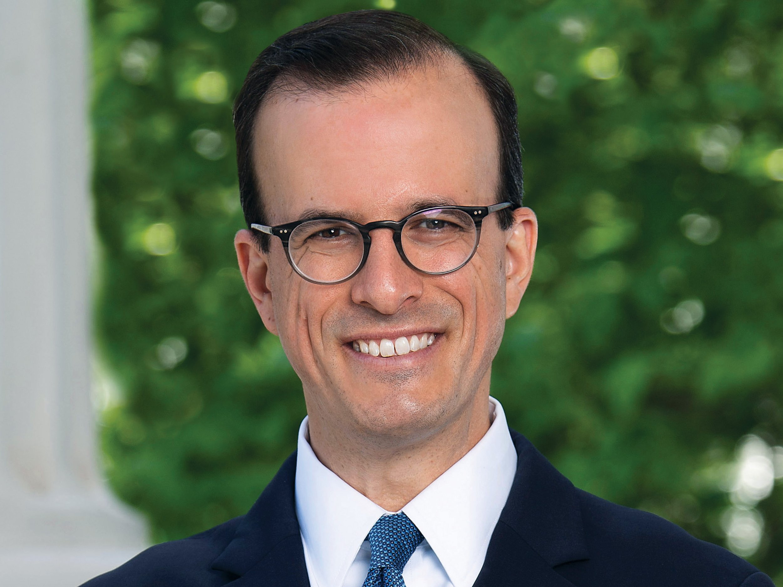 A man with short dark hair, round glasses, and a broad smile, dressed in a navy suit, white shirt, and blue tie, standing outdoors with green foliage in the background.