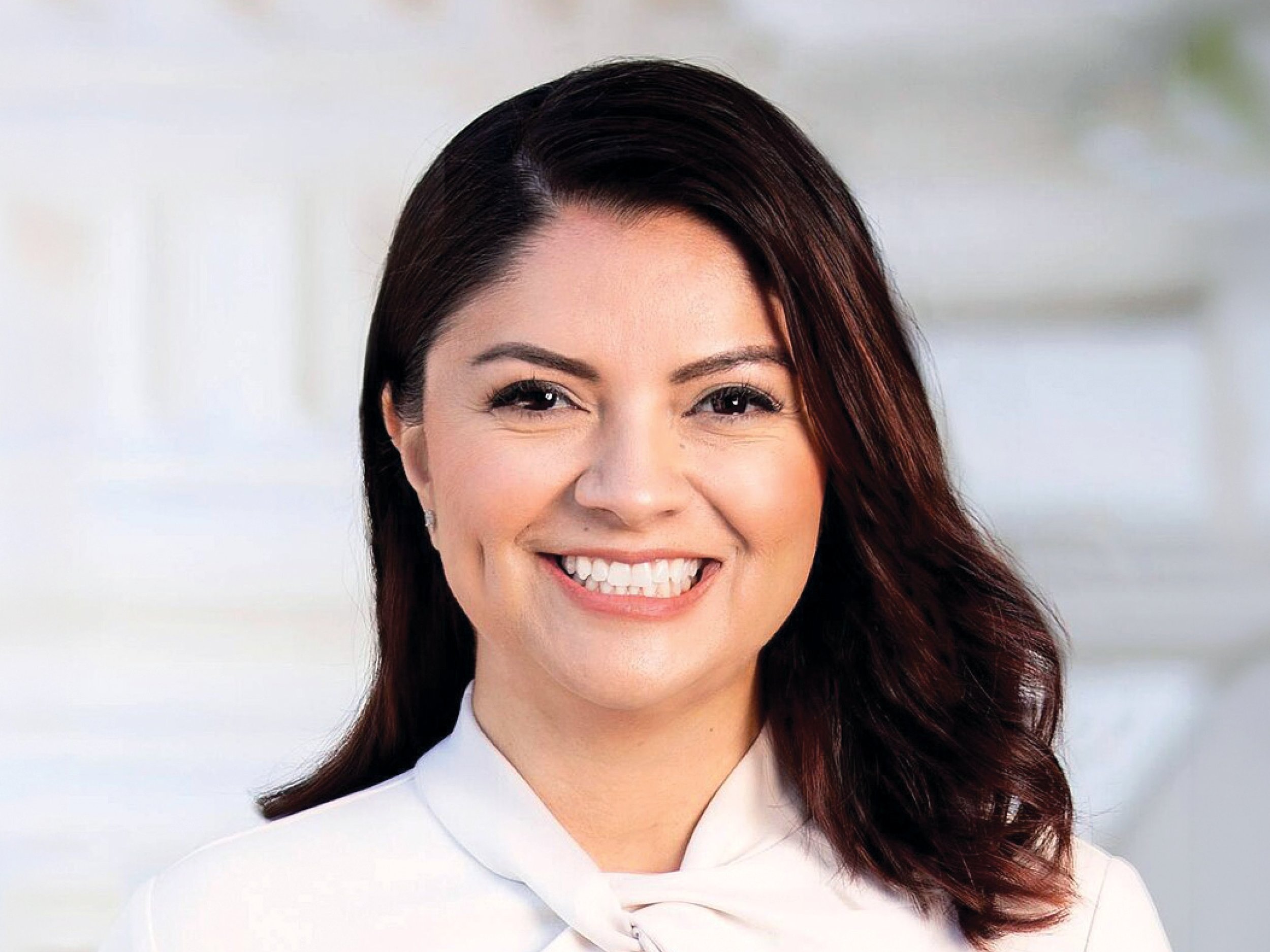 A woman with shoulder-length dark brown hair smiling, wearing a white professional outfit, in a bright, blurred office background.