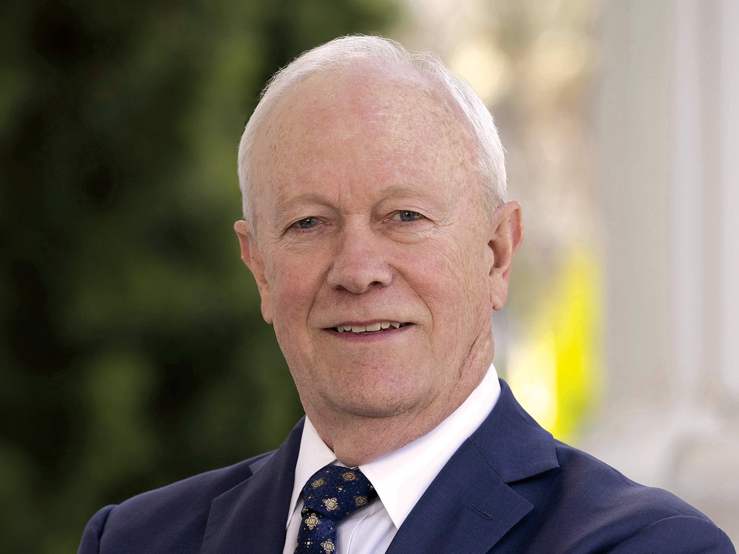 A portrait of an elderly man with light skin and short white hair, wearing a navy suit, white shirt, and patterned tie. He is smiling slightly and standing outdoors with a blurred background of greenery.