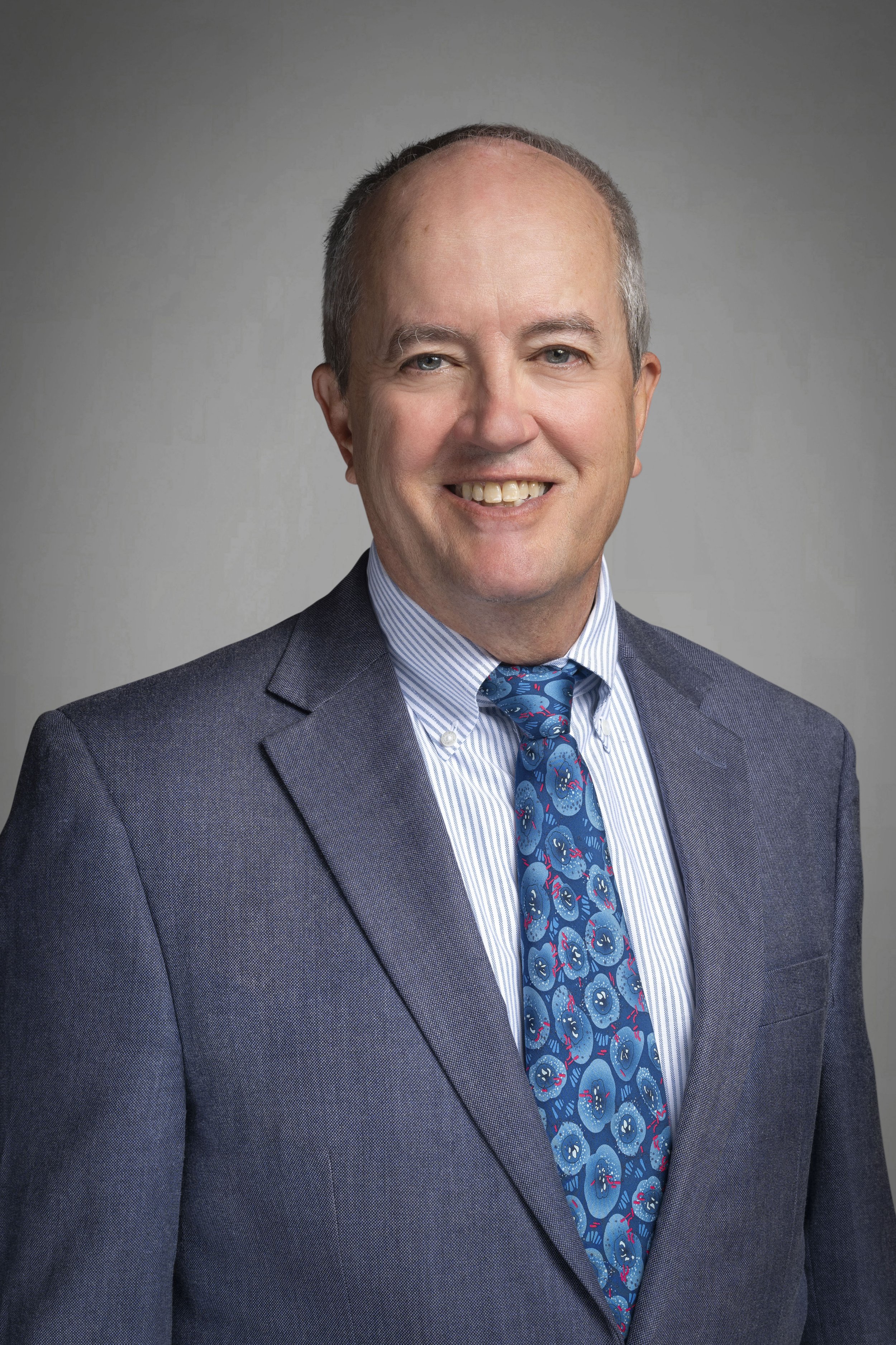 Headshot of a smiling middle-aged man wearing a dark blue suit, striped shirt, and a colorful blue tie with a pattern, against a neutral background.