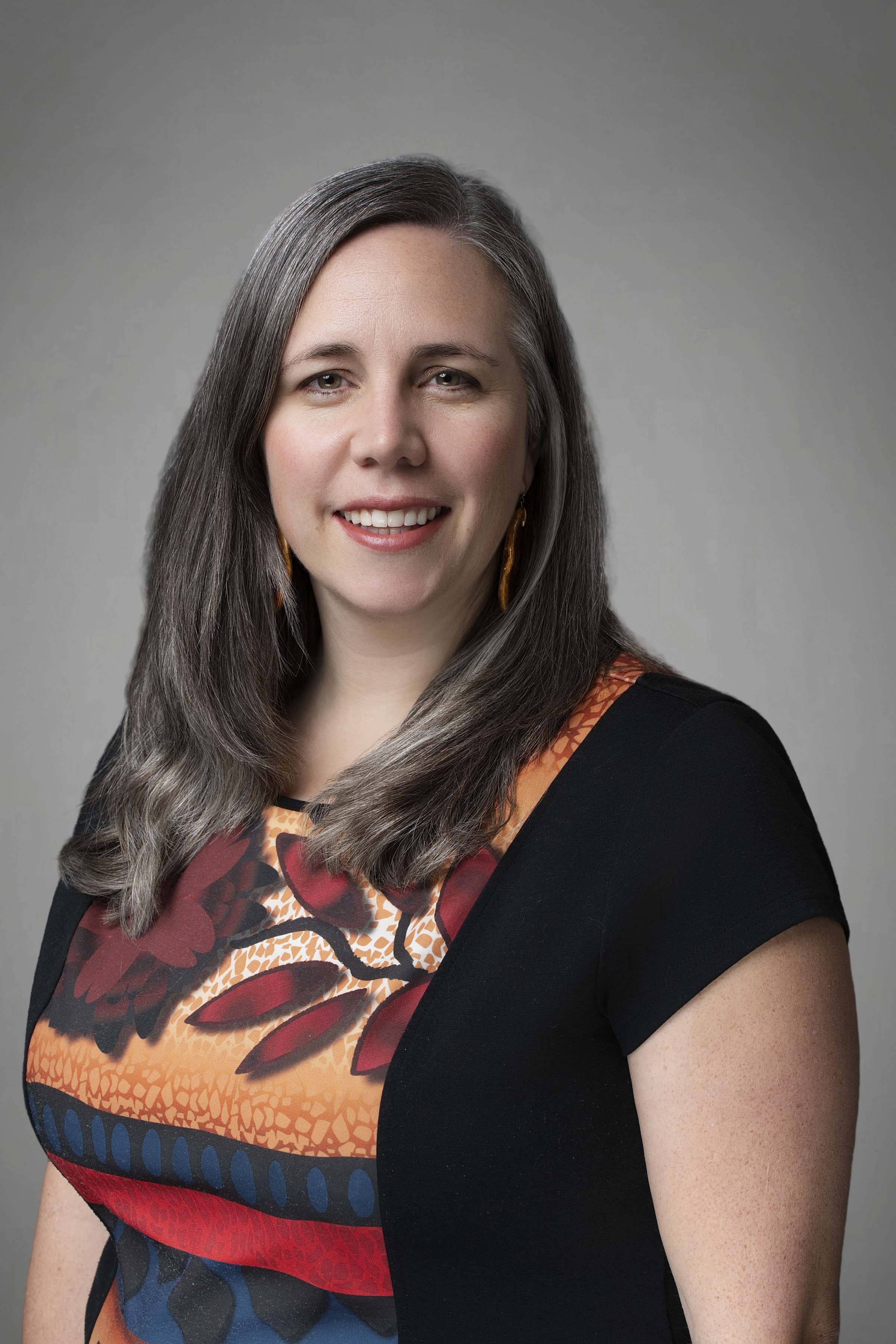 A woman with long, gray hair, smiling, wearing a colorful patterned dress and black top, with gold earrings, against a gray background.