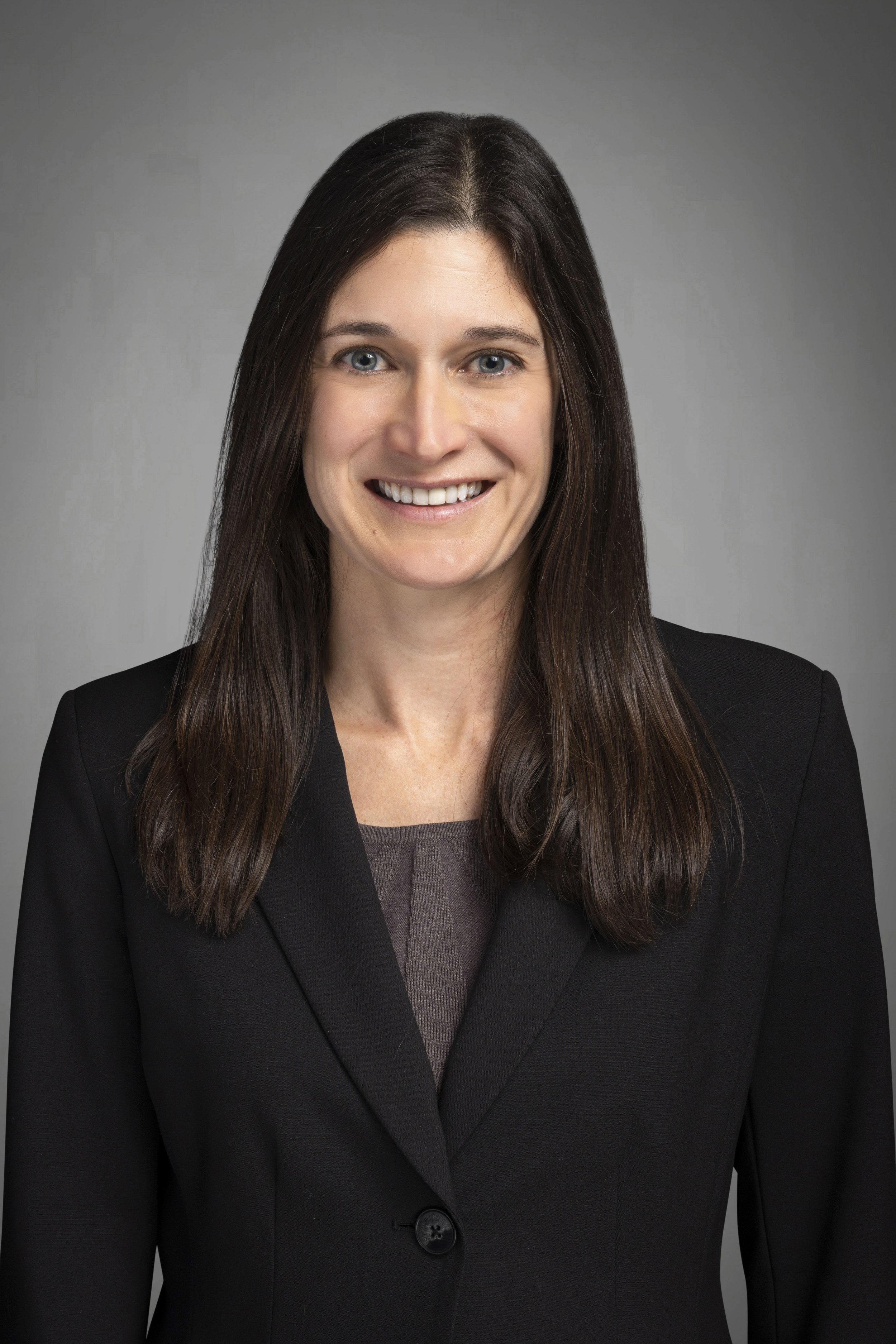 Headshot of a woman with long brown hair wearing a black blazer and gray top, smiling at the camera against a plain gray background.
