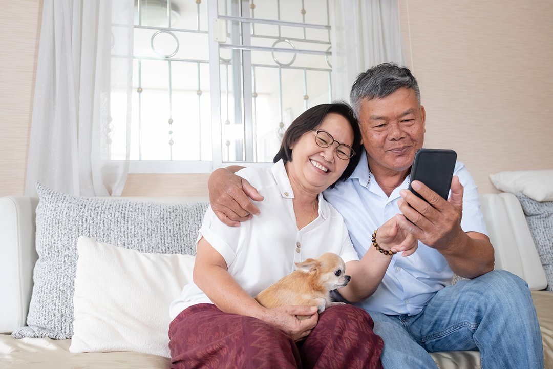 An elderly couple sitting on a couch, smiling at a smartphone, with a small dog in the woman's lap, in a cozy living room with curtains and pillows.