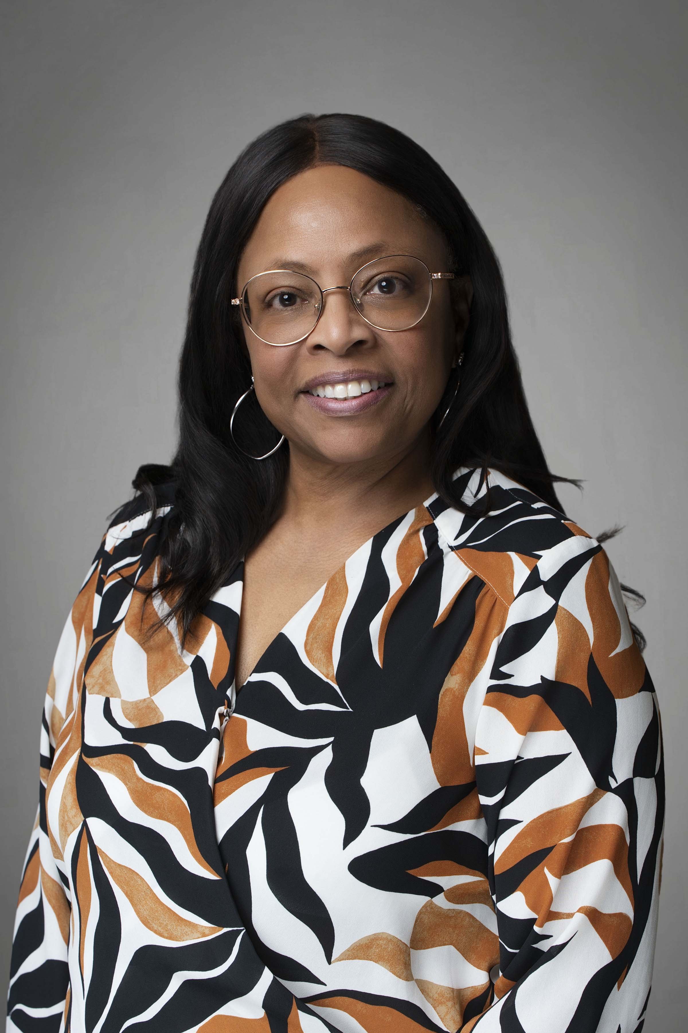 A woman with black hair, wearing glasses, hoop earrings, and a black, white, and brown leaf-patterned blouse, posing against a gray background.