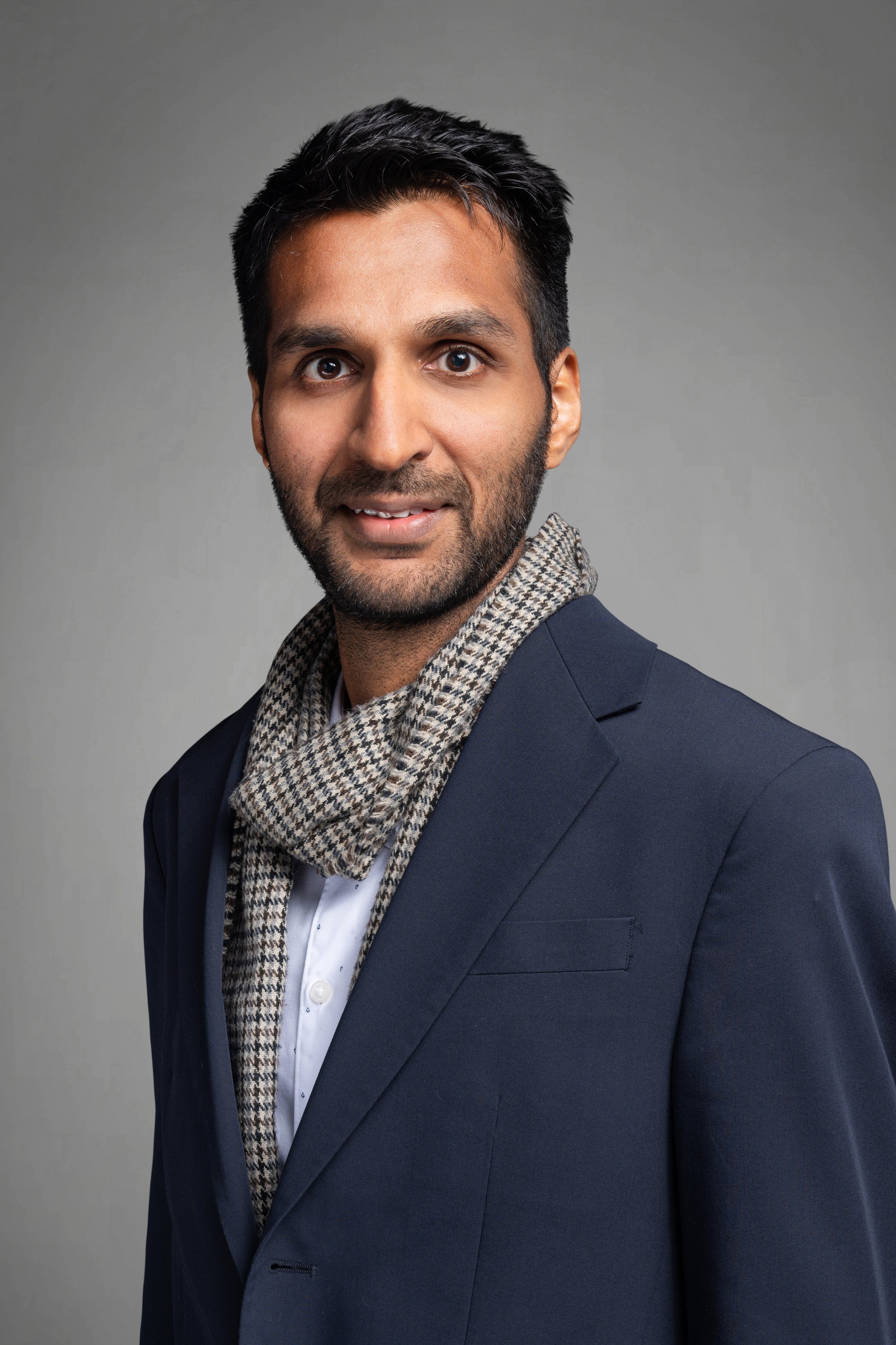 Professional portrait of a man with dark hair and beard wearing a navy suit, white shirt, and a checkered scarf against a gray background.