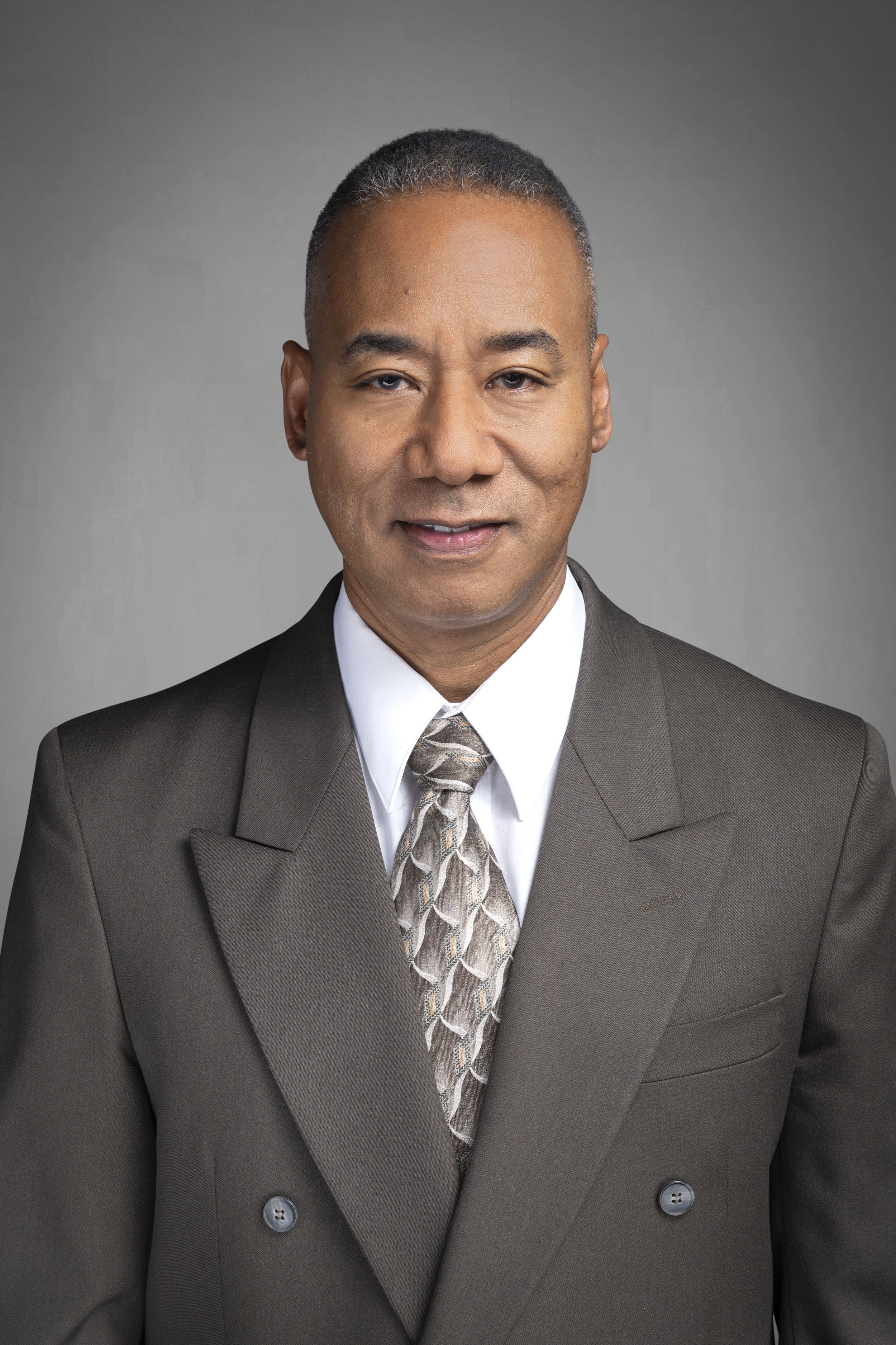 A professional headshot of a man in a brown suit, white shirt, and patterned tie, standing against a neutral gray background.