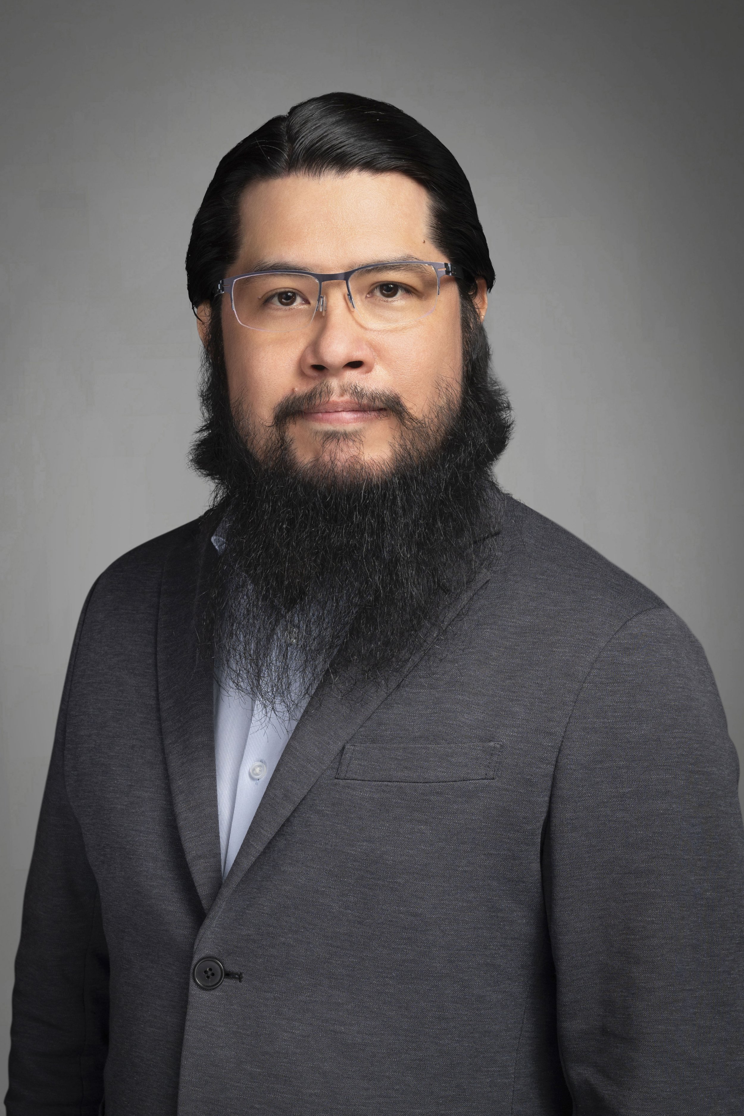 Portrait of an Asian man with black hair, glasses, a full beard, wearing a dark gray suit jacket over a light shirt against a gray background.