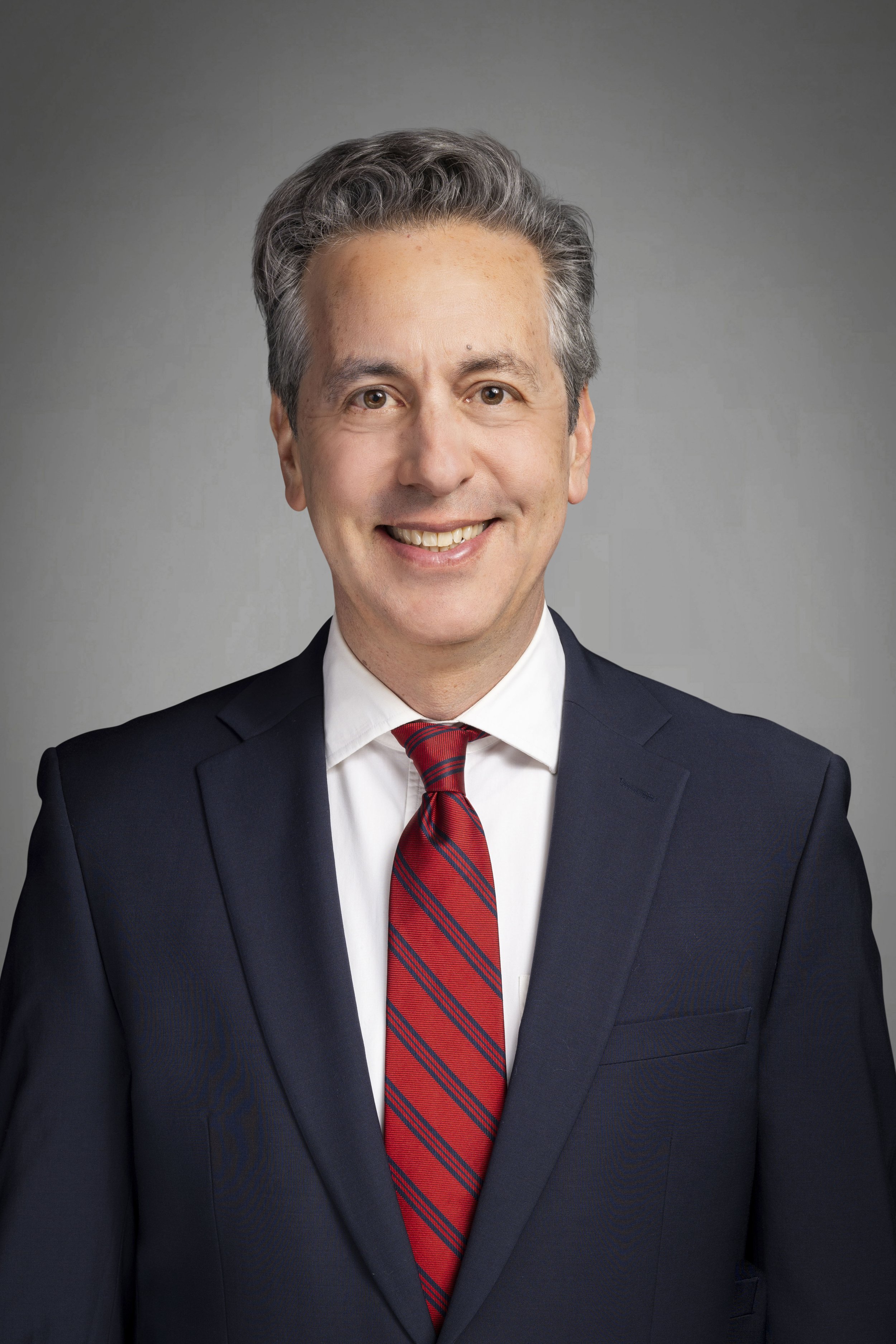 Professional portrait of a middle-aged man with gray hair, smiling, dressed in a dark suit, white shirt, and red striped tie, against a plain gray background.