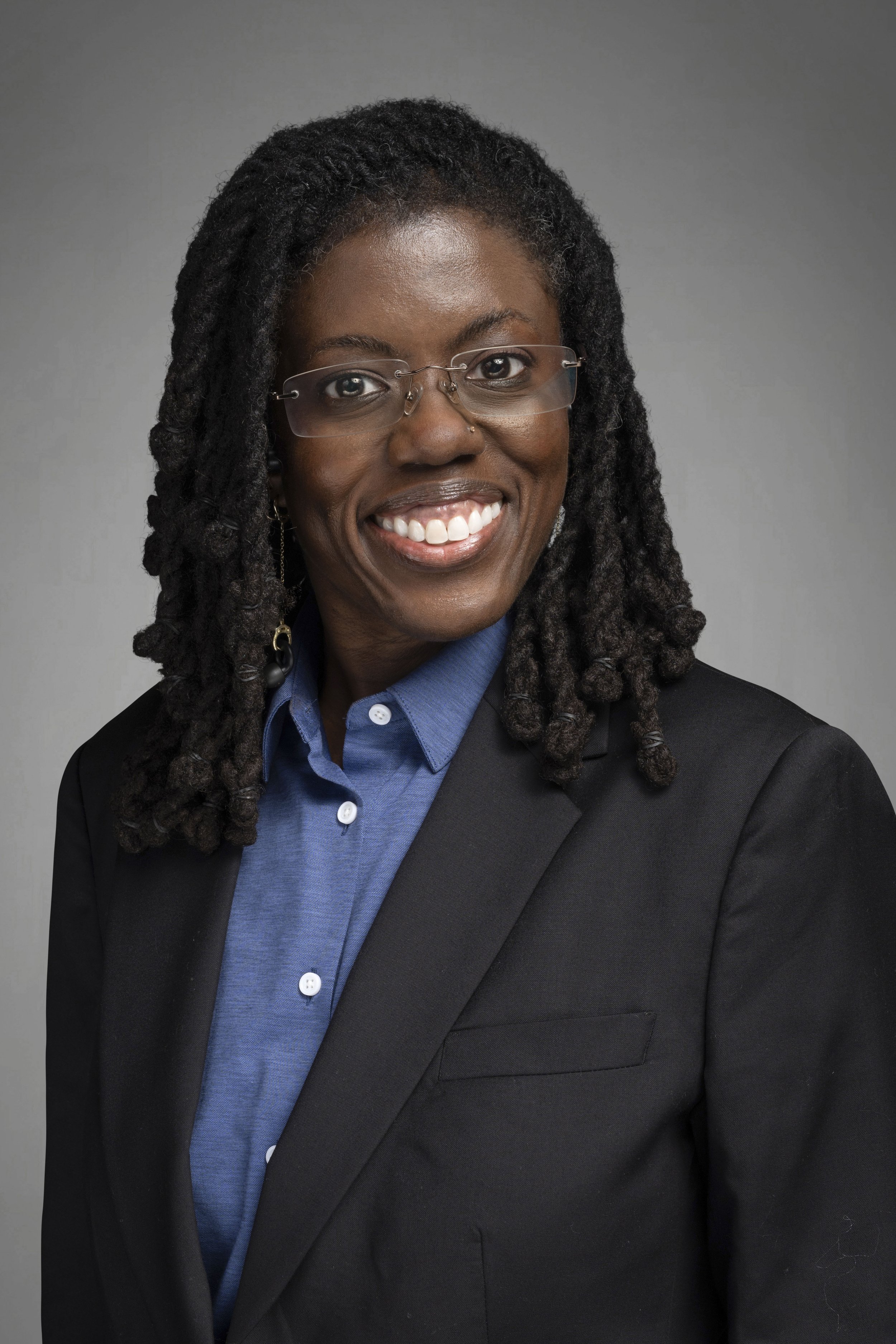 A woman with long dreadlocks, glasses, and a nose piercing, smiling while wearing a black blazer and a blue shirt against a gray background.