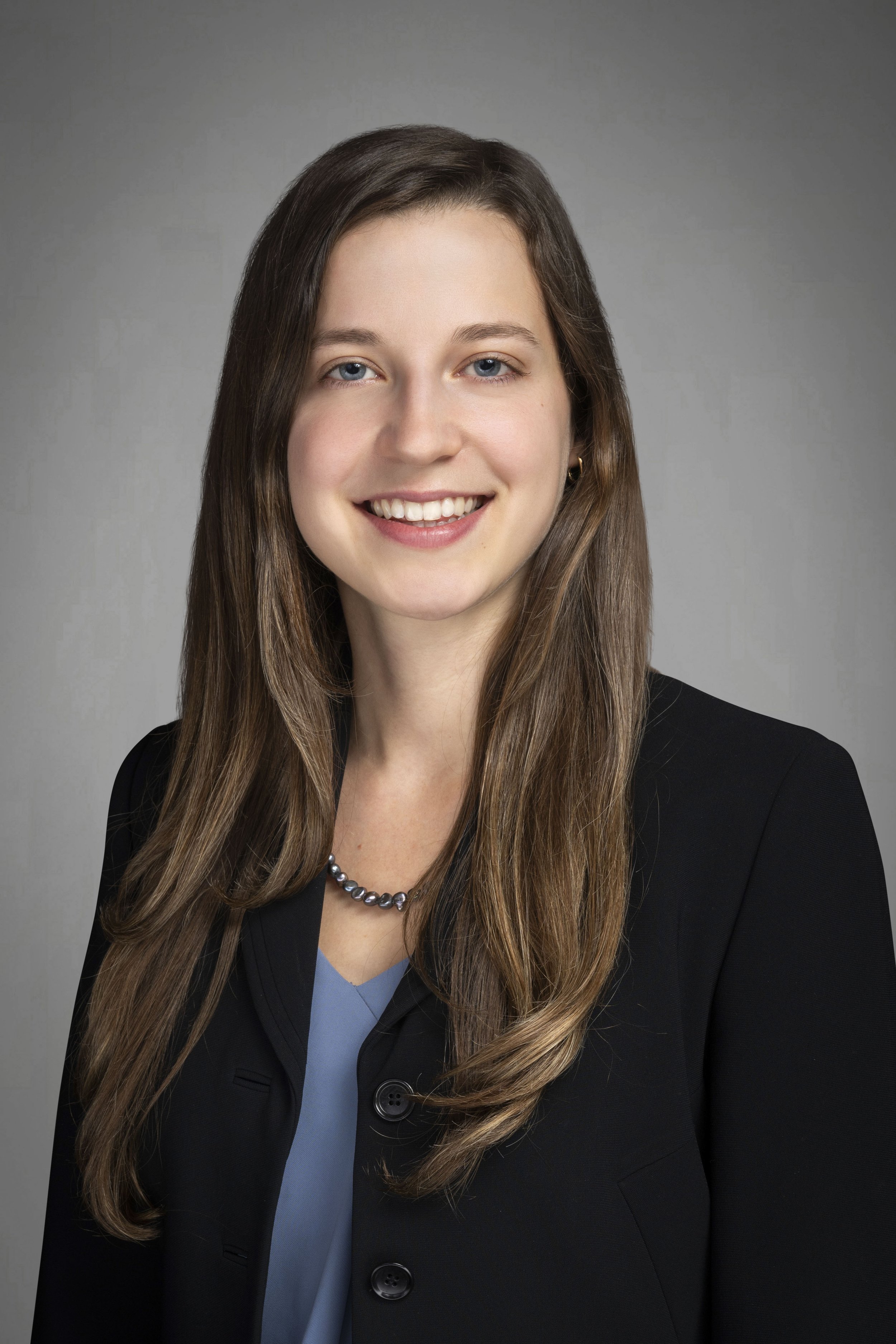 Professional headshot of a young woman with long brown hair, smiling, wearing a black blazer, a blue blouse, a pearl necklace, and small earrings, against a gray background.